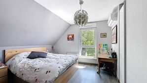 Een slaapkamer met een schuine kap, een bed met een houten hoofdeinde, een bureau met een stoel, en een groot raam.