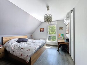Ein Schlafzimmer mit schrägen Wänden, einem Bett mit Holz-Kopfteil, einem Schreibtisch mit Stuhl und einem großen Fenster.