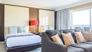 Habitación de hotel con cama, sofá, mesita de noche con lámpara y un amplio ventanal con cortinas.