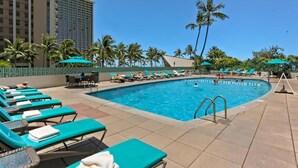 A pool area with lounge chairs and umbrellas, surrounded by palm trees and a tall building. - @ Marbella Lane - Luxe 2BR w/ Panoramic Ocean View (Honolulu)