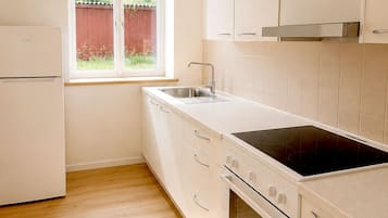 Una cocina con un refrigerador blanco, estufa, horno y pileta. Se observa una ventana que da a una cerca roja y vegetación al exterior.