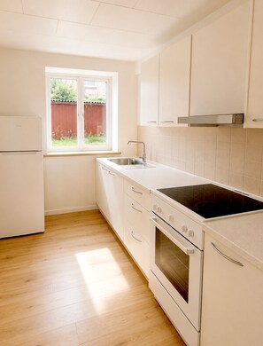 Una cucina con frigorifero, piano cottura, forno e lavandino bianchi. Si vede una finestra con vista su una recinzione rossa e del verde all'esterno.