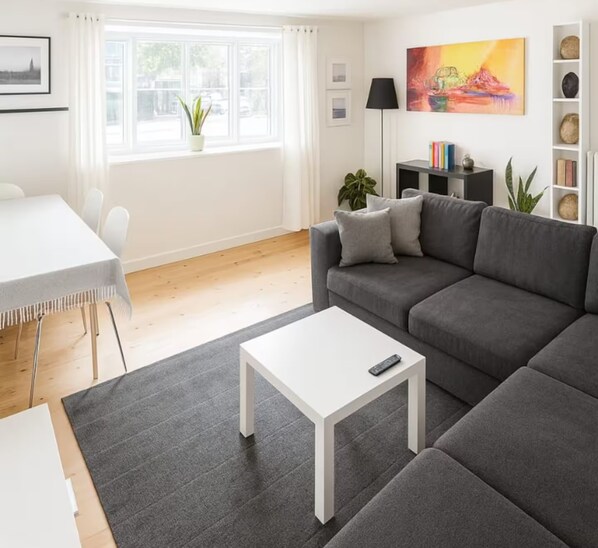 A modern living room with a grey sofa, a white coffee table, a dining area with a white table and chairs, and a large window with curtains.