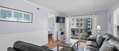 Un salon moderne avec un canapé en cuir, une table basse et un téléviseur. On aperçoit un balcon avec des meubles d’extérieur à travers les portes vitrées.
