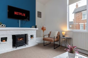 A living room with a fireplace, a television mounted on a blue wall, a comfortable chair, and a view of a brick building through a window. - 3BR Home in Darlington | Contractor Rates (Darlington)