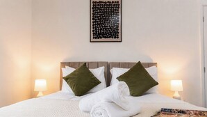 Een net opgemaakt bed met witte lakens en twee groene kussens, een ingelijste afbeelding boven het bed en aan beide zijden nachtlampjes.