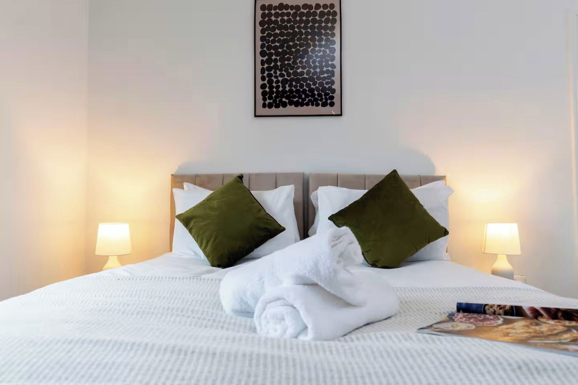 Una cama bien hecha con sábanas blancas y dos almohadas verdes, un cuadro enmarcado encima de la cama, y dos lámparas de noche.