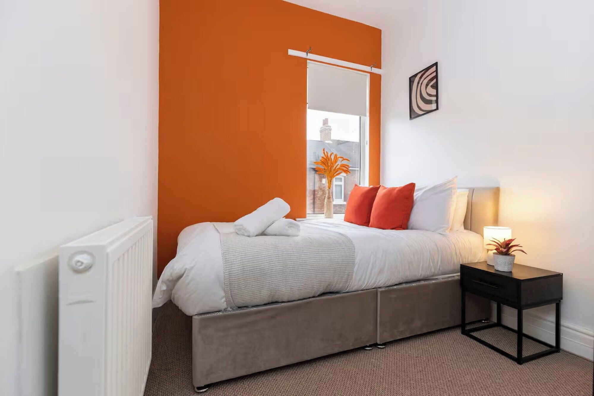 Et soveværelse med seng, natbord, radiator og et vindue med orange væg.
