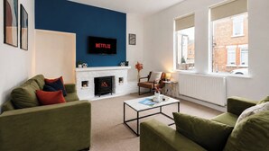 Een woonkamer met een open haard, groene banken, een salontafel en een televisie bevestigd aan een blauwe muur.