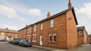 Een herenhuis met twee verdiepingen, een garage, geparkeerde auto's en een straat met kinderkopjes.