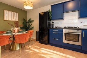A modern kitchen with a black refrigerator, blue cabinets, and a dining area with orange chairs and a table set for four. - Darlington 3BR – Bright, Spacious & Stylish (Darlington)