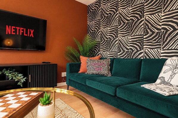 A living room with a green sofa, a flat-screen TV mounted on the wall, a patterned wall, and a small plant on a side table. - Darlington 3BR – Bright, Spacious & Stylish (Darlington)