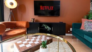 A living room with a television displaying 'NETFLIX,' a glass coffee table with a chessboard, a floor lamp, and a green sofa. - Darlington 3BR – Bright, Spacious & Stylish (Darlington)