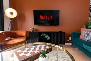 Ein Wohnzimmer mit einem Fernseher, der 'NETFLIX' anzeigt, einem Glastisch mit Schachbrett, einer Stehlampe und einem grünen Sofa.