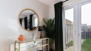 A room with a glass console table, a round mirror with a gold frame, a potted plant, and a view through a sliding glass door. - Darlington 3BR – Bright, Spacious & Stylish (Darlington)