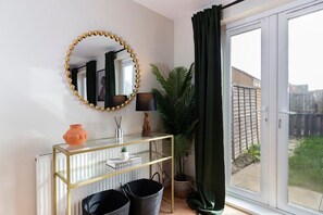 Ein Zimmer mit einem Glastischchen, einem runden Spiegel mit goldener Rahmung, einer Topfpflanze und Blick durch eine Glastür.