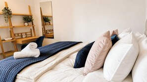 Una cama bien hecha con colcha azul, almohadas blancas y una toalla blanca enrollada. Hay una estantería de madera con plantas y un reloj, y un espejo en la pared.