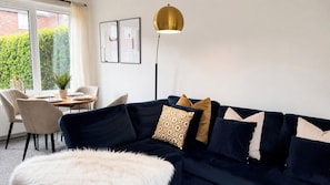 Ein Wohnzimmer mit einem dunkelblauen Sofa, einem weißen, flauschigen Ottomanen, einem Tisch mit Stühlen und einer Stehlampe.