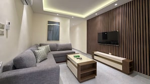 A modern living room with a sectional sofa, a flat-screen TV mounted on a wooden panel wall, and recessed ceiling lighting. - 106 JRE One-br Apartment in Amman (Amman)