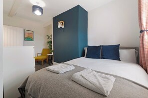 Ein Hotelzimmer mit Bett, zwei Kissen, Kopfteil, Nachttisch, Sessel, Pflanze und Wanddeko.
