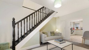Eine Treppe mit schwarzen Spindeln, ein Wohnzimmer mit einer Couch und einem Couchtisch sowie eine Küche im Hintergrund.