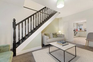 Eine Treppe mit schwarzen Spindeln, ein Wohnzimmer mit einer Couch und einem Couchtisch sowie eine Küche im Hintergrund.