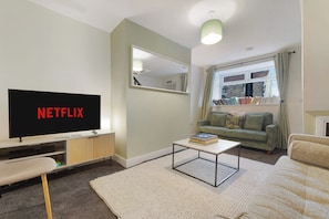 Ein Wohnzimmer mit Fernseher, der 'NETFLIX' zeigt, einer Couch, einem Couchtisch und einem Fenster mit Vorhängen.
