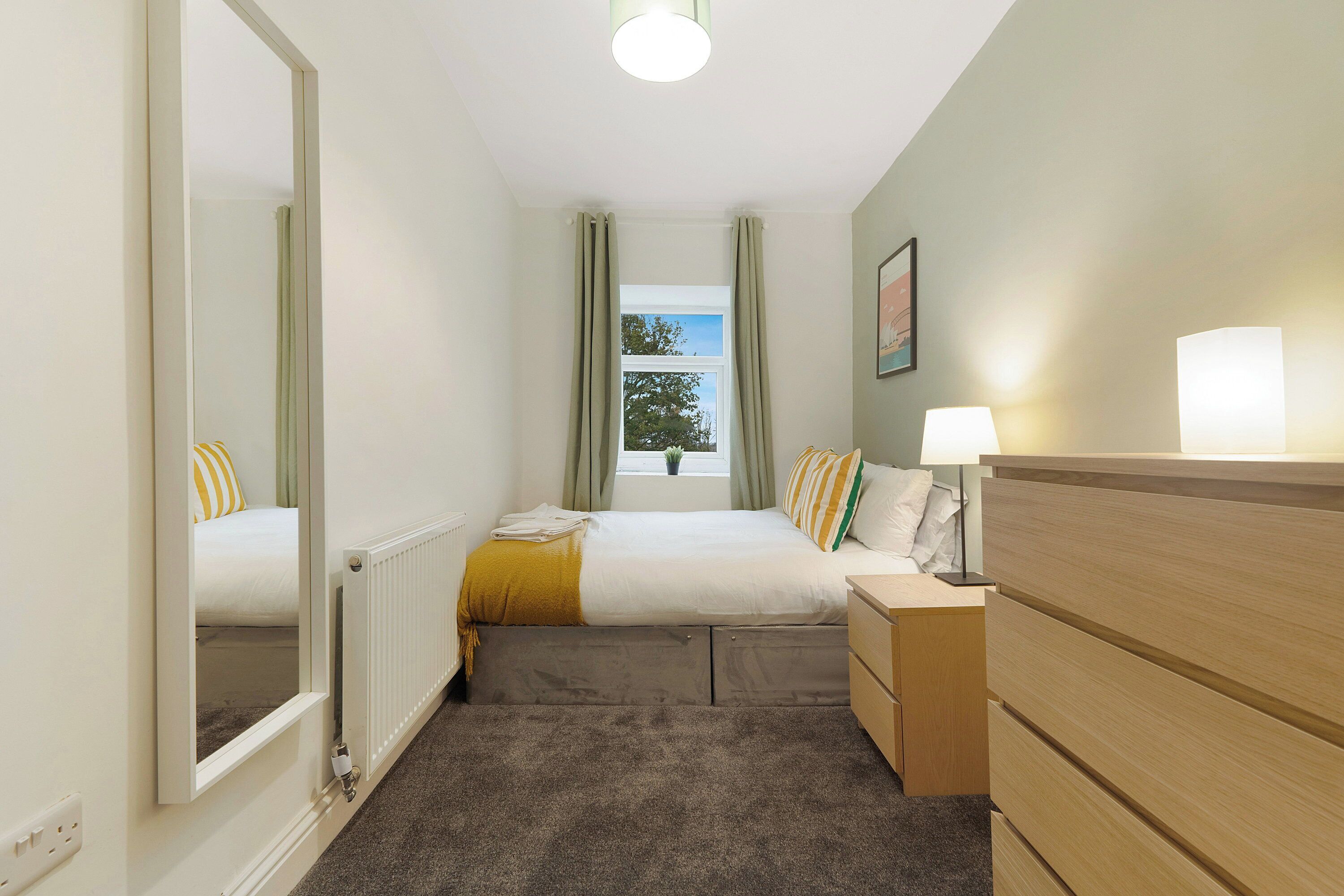 Une chambre à coucher comprenant un lit, une commode, un miroir, une fenêtre avec des rideaux et un radiateur.