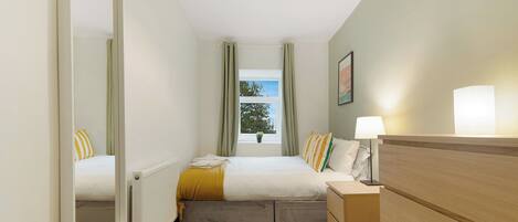 A bedroom with a bed, a dresser, a mirror, a window with curtains, and a radiator.