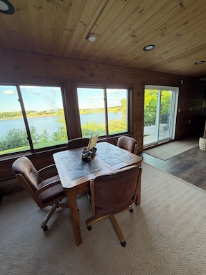 Un intĂ©rieur en bois avec une grande table et des chaises, de grandes fenĂȘtres donnant sur un lac et la verdure, et un tapis Ă motifs en chevrons.