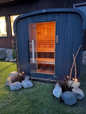 Eine Holzsauna mit Glastür, umgeben von Steinen und Pflanzen, und einer dunklen Außenwand.