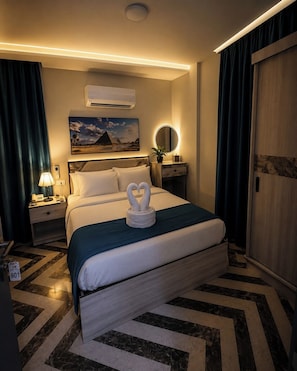 A hotel room with a bed, nightstand, lamp, air conditioning unit, and a painting on the wall.