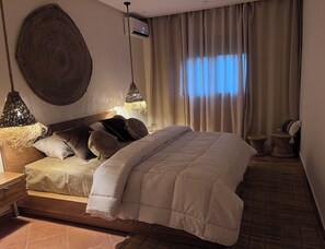 Ett sovrum med en säng, sängbord, en hängande lampa, en väggmonterad luftkonditioneringsanläggning och ett fönster med gardiner.