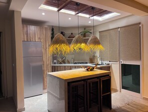 Eine moderne Küche mit Marmorarbeitsplatte, Holzschränken, einem integrierten Kühlschrank, einer hängenden Pendelleuchte und einem Fenster mit Jalousien.