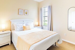 Une chambre à coucher bien rangée, avec un lit, des tables de chevet, une fenêtre avec des rideaux et des tableaux encadrés accrochés au mur.