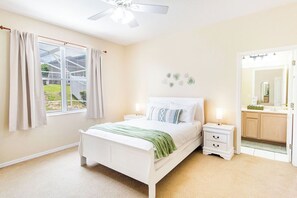 Ein Schlafzimmer mit Bett, Nachttischen, Deckenventilator, Fenster mit Vorhängen, Spiegel und Blick ins Freie.