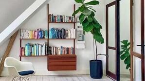 Ein Loft-Zimmer mit schrägem Dach, Holzbalken, einem Bücherregal, einer Topfpflanze, einem weißen Stuhl und einer Schiebetür aus Glas.