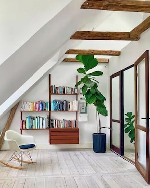 Ein Loft-Zimmer mit schrägem Dach, Holzbalken, einem Bücherregal, einer Topfpflanze, einem weißen Stuhl und einer Schiebetür aus Glas.