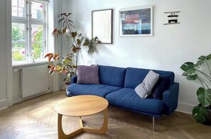 A living room with a blue sofa, a round wooden coffee table, and a large window with a view of greenery. - 1881 townhouse in Kartoffelrækkerne near the Lakes (København Ø)