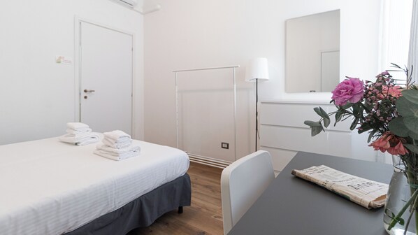 Quarto de hotel com uma cama, uma mesa com um jornal, um vaso com flores e um espelho.