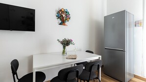Een moderne keuken met een witte tafel, zwarte stoelen, een koelkast, een televisie en een decoratieve boom.