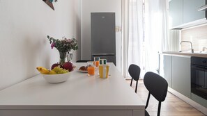 Uma cozinha moderna com bancada branca, geladeira e área de refeições com cadeiras e mesa posta para o café da manhã.