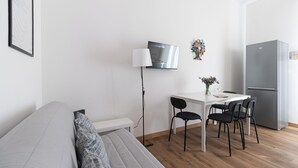 Een moderne, minimalistische kamer met een bank, een eettafel met stoelen, een flatscreen televisie, een koelkast en een vloerlamp.