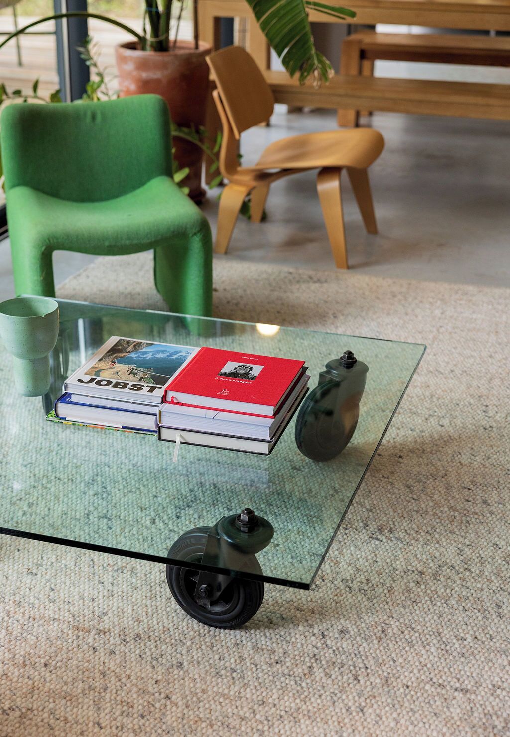 A glass table with a stack of books, a small vehicle, and a green chair in a room with a carpeted floor.