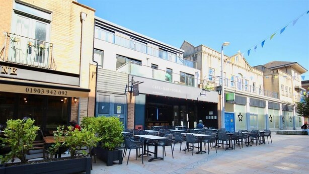 Ein Straßen-Café mit Tischen und Stühlen, ein Gebäude mit Schild "Café-Bar-Sozial", und ein klarer Himmel.