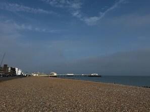 Ein Kiesstrand mit einem Steg, der sich ins Wasser erstreckt, Gebäuden im Hintergrund und einem klaren Himmel.