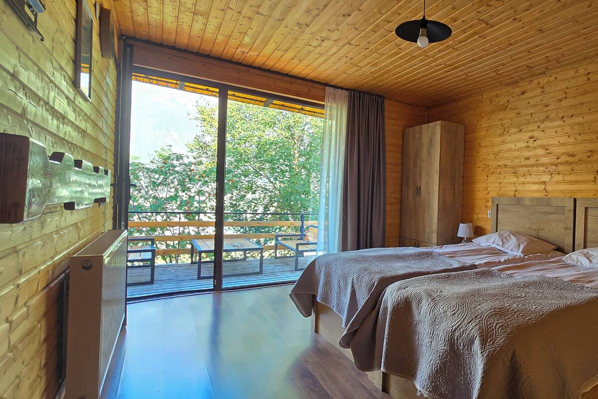 A wooden cabin bedroom with two beds, a balcony, and a view of trees.