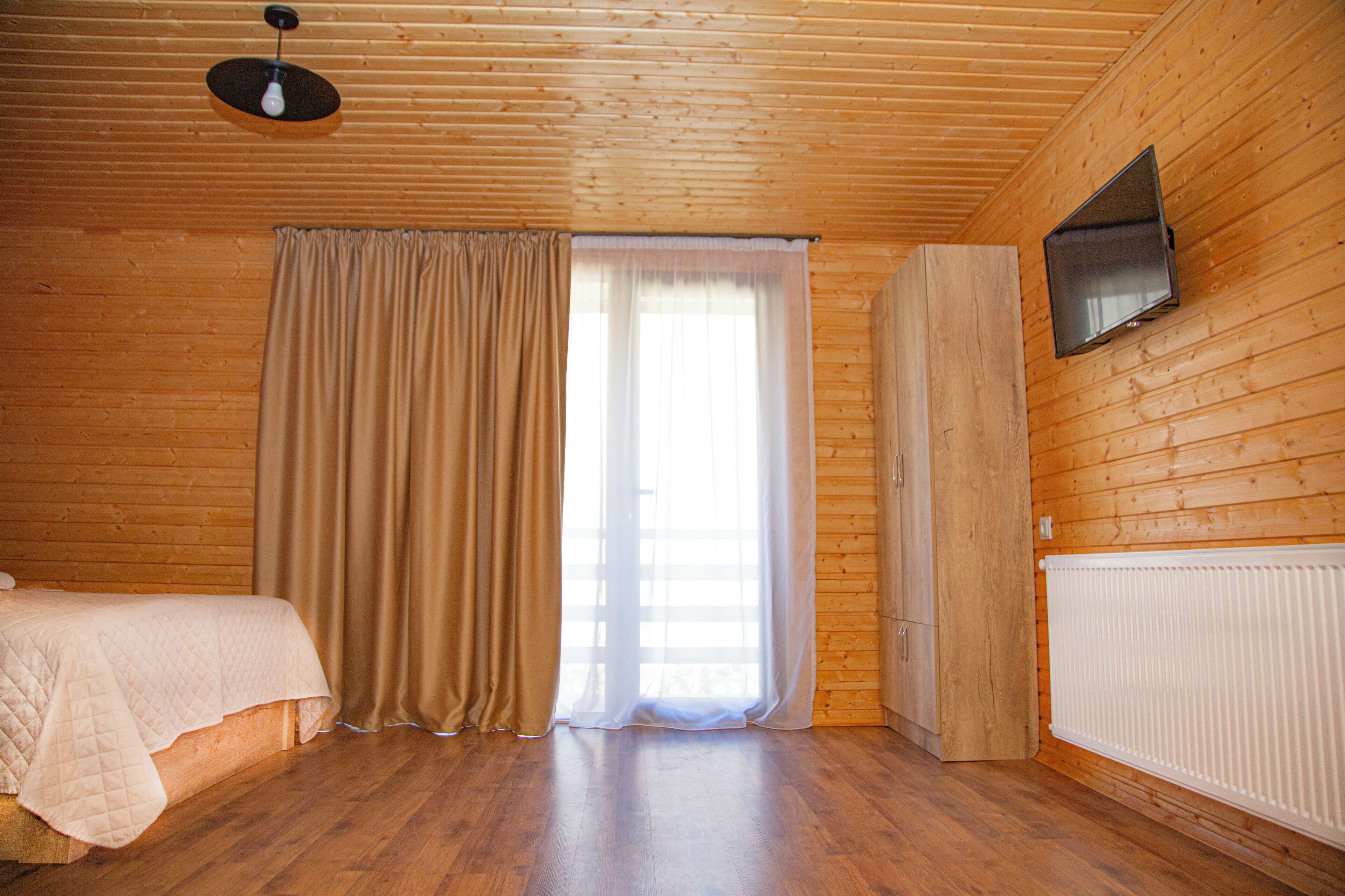 A room with wooden walls, a bed with a quilted cover, a window with curtains, a radiator, and a mounted television.