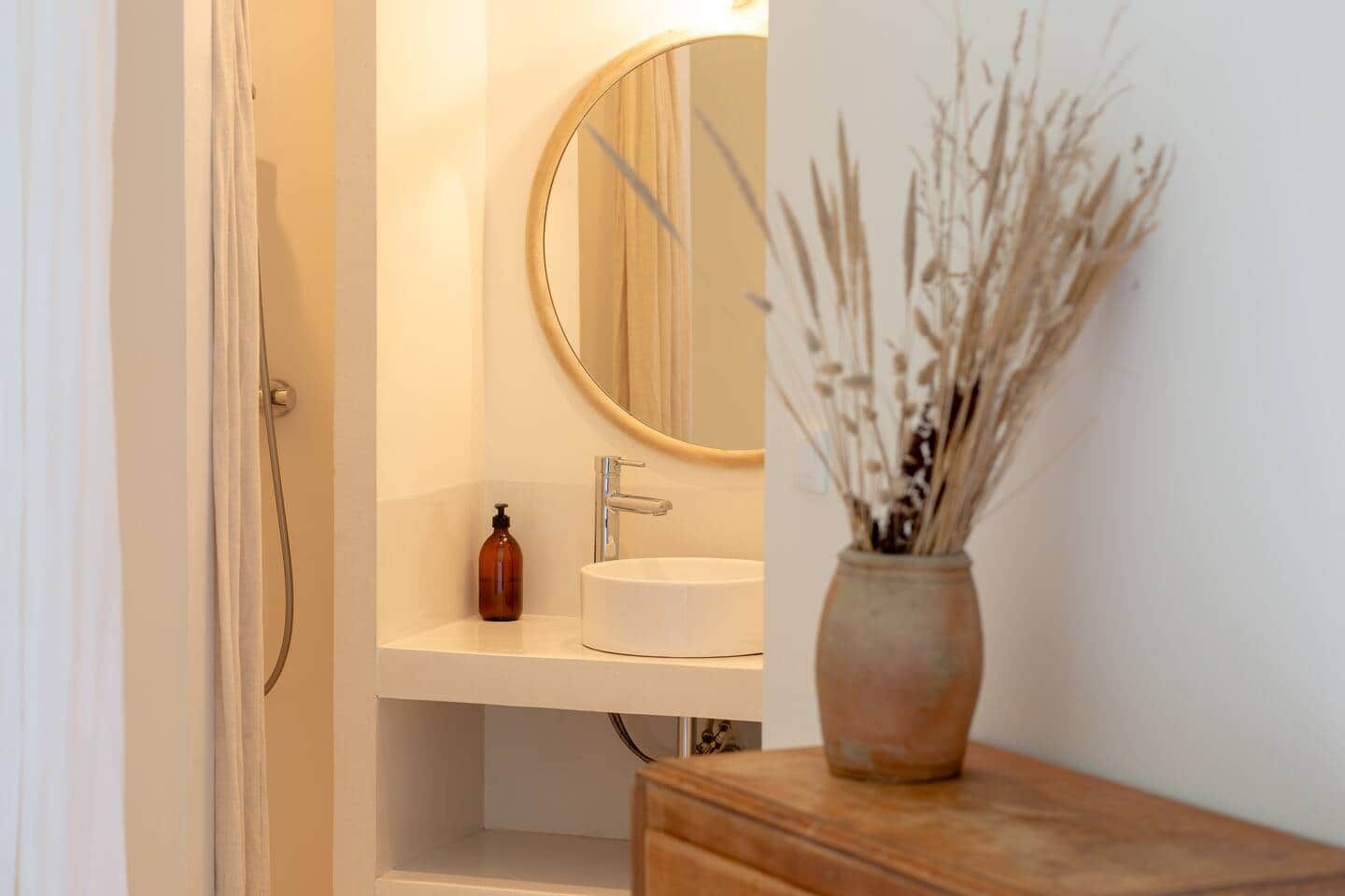 Une salle de bain avec un lavabo blanc, un miroir rond, un meuble-lavabo en bois et un vase contenant des plantes séchées.