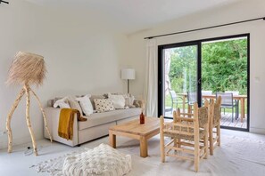 Ein modernes Wohnzimmer mit Sofa, Couchtisch und Sesseln, einer Stehlampe und Blick auf einen Garten durch eine Glasschiebetür.